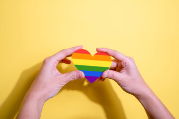 Rainbow heart shape in female hand on yellow background. LGBT, Pride, LGBTQ Symbols