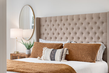 A bedroom detail shot with a cushioned headboard, a gold mirror on the wall, and a plant and lamp...