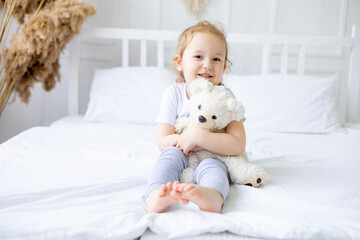 a little cute baby girl on a white cotton bed at home with a soft toy in her hands smiling on the bed at home