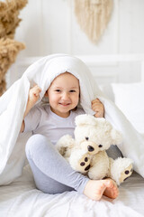 a little cute baby girl peeks out from under the blanket on a white cotton bed at home smiling hugging a teddy bear, the child plays and indulges at home in the morning