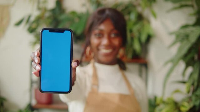 Forist African American woman holding phone with chroma key. Greenhouse worker female botanist smiling, small business owner. Gardener working in flower shop, plant store. 