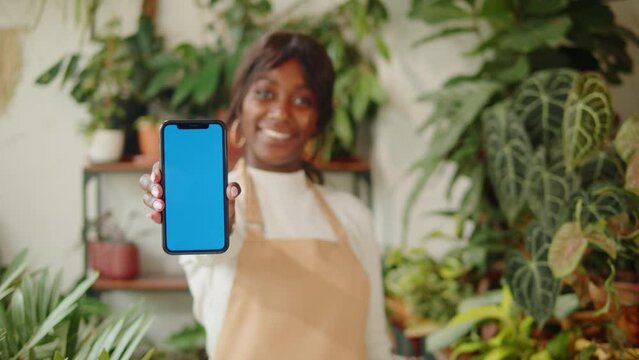 Forist African American woman holding phone with chroma key. Greenhouse worker female botanist smiling, small business owner. Gardener working in flower shop, plant store. 