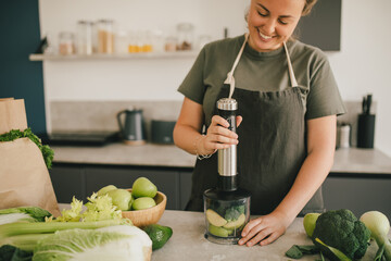 Young woman making smoothie at the kitchen, using blender and fresh fruits and vegetables.