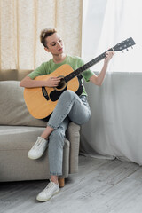 full length view of young pansexual person in jeans playing acoustic guitar on sofa.