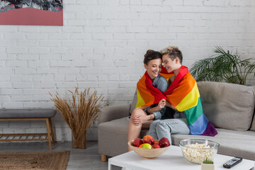 happy pangender couple sitting on couch under lgbt flag near apples and popcorn on coffee table.