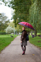 Young smilling short haired blonde woman walks in the autumn park with red umbrella. View from back.