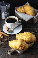 Fresh crisp crunchies lie on a wooden plank. A white cup with black coffee and a coffee pot in the background. Coffee beans and cinnamon straws on the table. Dark background