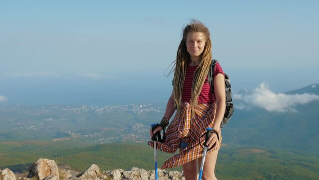 Young woman with dreadlocks is practicing Nordic walking. Sports girl with backpack and poles stands on mountain. Female tourist enjoys views of nature on top of hill