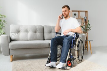 The disabled person sits in a wheelchair. He is talking to someone on his smartphone. He is in his large bright living room. He smiles.