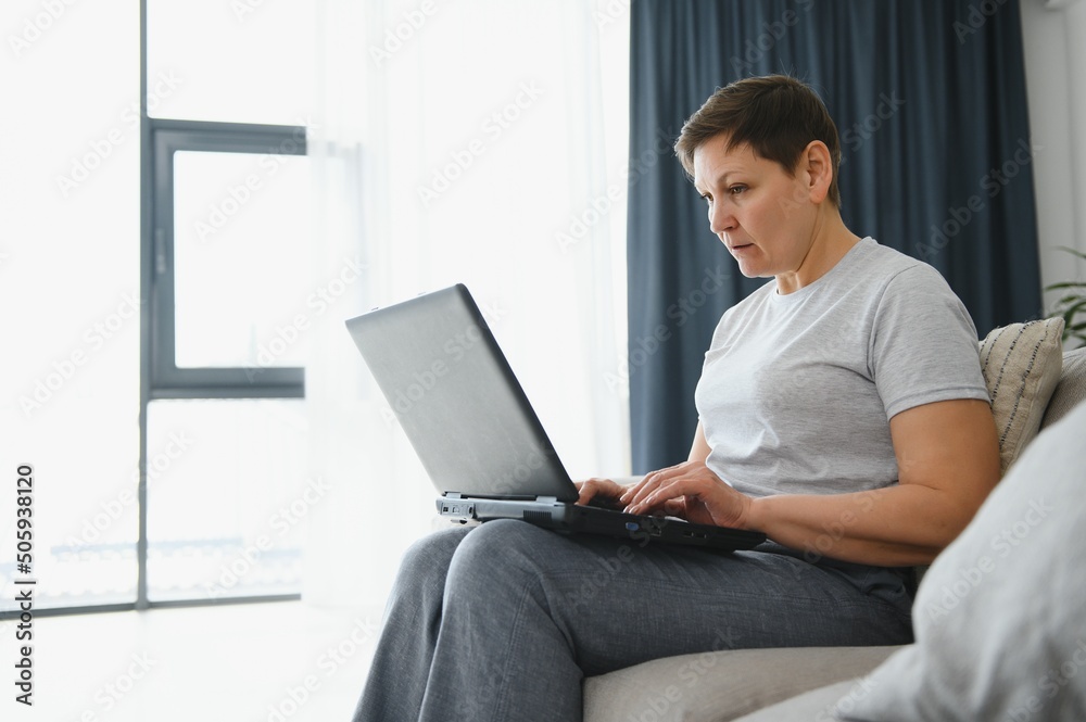 Poster Excited mature woman looking at laptop screen, reading good news in message, celebrating online lottery win, rejoicing success, overjoyed older female sitting on couch at home, using computer.