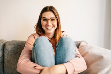 Young woman with glasses looks at camera laughing and sitting comfortably on sofa