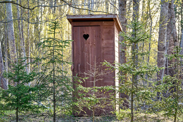 Wooden outdoor toilet with heart on the door. Spruce trees around.