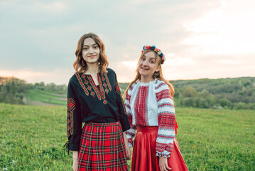 two girls in traditional ukrainian clothes - vysyvanka holding hands and smiling at camera in the field during sunset. 
