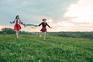two girls in traditional ukrainian vysyvanka holding ukrainian flag and running in the field during sunset. Symbol of bravery and independence. Stand with Ukraine