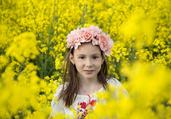 Face portrait of a cute girl 7 years old in a wreath and a traditional embroidered blouse among a yellow flowering rapeseed field. Children for Peace. Stand with Ukraine. Be proud to be Ukrainian