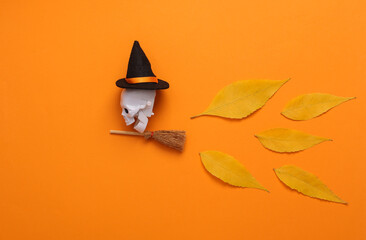 Fototapeta premium Skeleton skull on a witch's broom with hat on orange background with fallen autumn leaves. October 31st, happy halloween concept, minimal layout