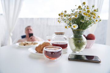 A glass teapot, a cup of chamomile tea and a smartphone on the background of a vase with daisies. In the background, a blurry man is looking at a smartphone. High quality photo
