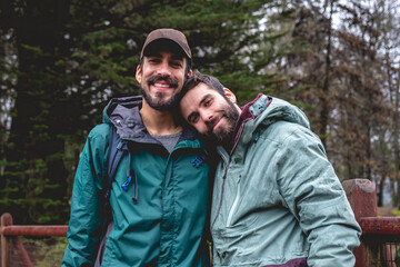 Two happy, young, caucasian, bearded and handsome brothers with jacket, cap and backpacks in a wooden bridge in the forest in a rainy day, Valdivia, Chile