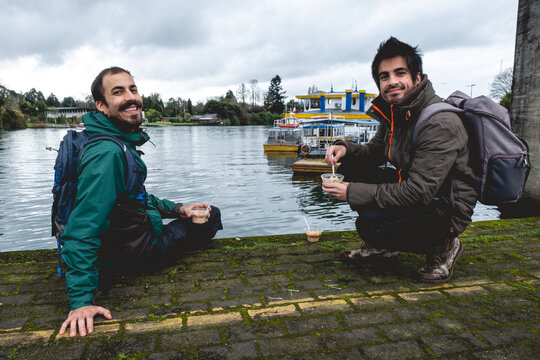 Two young, bearded, happy and handsome brothers with jacket and backpack eating ceviche (fresh raw fish with citrus juice and chili peppers) and panoramic view of river, boats and cloudy sky, Valdivia