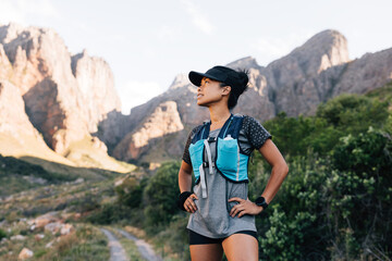 Naklejka premium Trail runner in cap relaxing in valley. Sportswoman looking away and smiling preparing for hike.