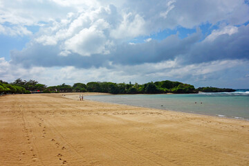 empty tropical beach, cloudy sky, yellow sand near blue water, trees in forest in distance, traces on sand 