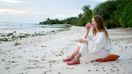 Fototapeta na wymiar woman in white bathrobe sitting on sand at beach, drinking cup of hot drink, enjoy morning near ocean, connect with nature
