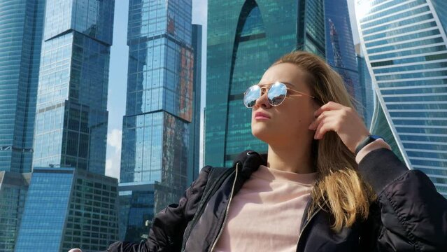 Portrait of beautiful blonde girl in sunglasses. Pretty lady posing against of skyscrapers with mirrored walls. Young woman admiring views of metropolis near glass high rises. Daytime