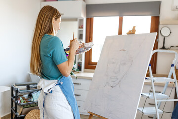 Young awesome cheerful girl enjoying painting on canvas at home. Portrait of beautiful woman paint in her art home studio. Young girl holding her paintbrush. Artist workshop education concept.