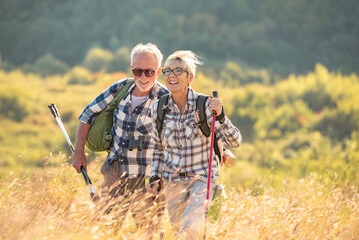 Active senior Caucasian couple hiking in mountains with backpacks and hiking poles, enjoying their adventure - obrazy, fototapety, plakaty