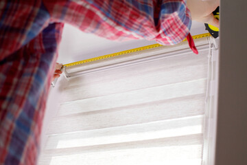 A handywoman with a tape measure installs white roller blinds indoors. Shutters day and night.