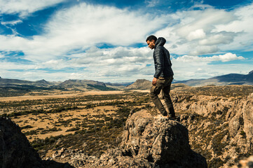 Young man standing on a rock at the top of a cliff overlooking a valley with a mountain range in the background. Concept of overcoming. Argentinian Patagonia - obrazy, fototapety, plakaty