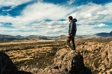 Young man standing on a rock at the top of a cliff overlooking a valley with a mountain range in the background. Concept of overcoming. Argentinian Patagonia - obrazy, fototapety, plakaty