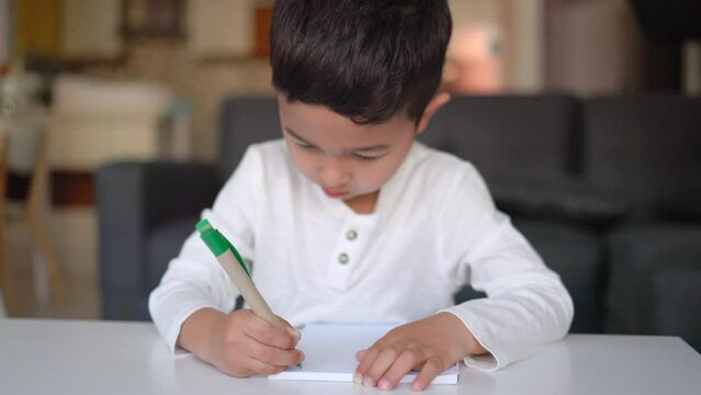 Little asian boy in white writing with blue pencil on paper sitting at home
