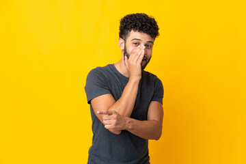 Young Moroccan man isolated on yellow background pointing to the side to present a product and whispering something - obrazy, fototapety, plakaty