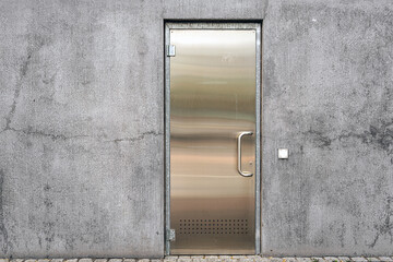 Shiny steel door with a metal handle and a lock on a gray old concrete wall next to a white electrical socket outdoors, minimalistic background