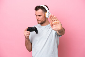 Fototapeta na wymiar Young Brazilian man playing with video game controller isolated on pink background making stop gesture and disappointed