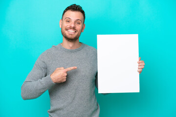 Young Brazilian man holding an empty placard isolated on blue background holding an empty placard with happy expression and pointing it