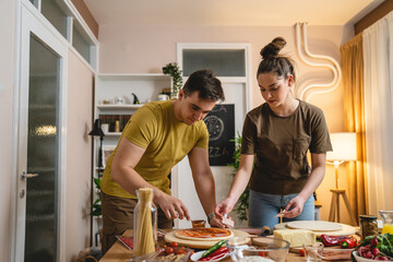 Two people young adult couple man and woman husband and wife or boyfriend and girlfriend preparing pizza food at home helping each other cutting and add ingredients and spices real people copy space