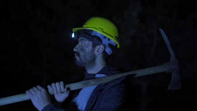 Tired miner looking at camera.
Mandeci looks around with the light of a lantern in the dark. He has a pickaxe. He has black dirt on his face.
