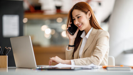 Asian businesswomen have the joy of talking on the phone with a laptop computer tablet on the office desk. doing accountant on a calculator to calculate business data documents.