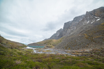 Atmospheric landscape with azure mountain lake in green valley and rocky mountain range with peaked top in low clouds. Beautiful alpine lake and high sharp rocks under cloudy sky at gloomy weather.