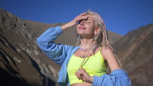 Portrait of joyful happy young woman with white dreadlocks in colourfull tank top. standing alone at background of Alp mountains and forest.
