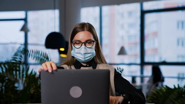 Focused young female employee in mask works at computer. Woman wearing glasses closes her laptop and looks straight into camera.