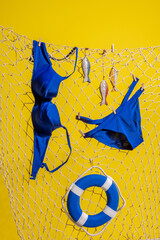 A blue swimsuit and wooden fish with a lifebuoy hang on a fishing net on a yellow background