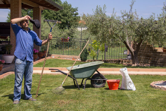 Image of a tired gardener who wipes sweat from his forehead in the garden at home while with a rake he cleans the lawn of leaves and dry grass.