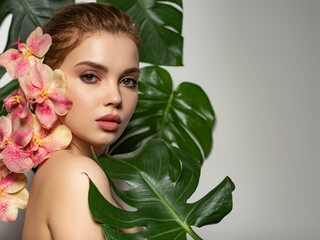 Obraz na płótnie Canvas Model with pink tropical orchid near face. Closeup girl's face with green leave. Pretty girl in Skin care beauty treatments concept. ​Beautiful woman with green leave near face and body. 