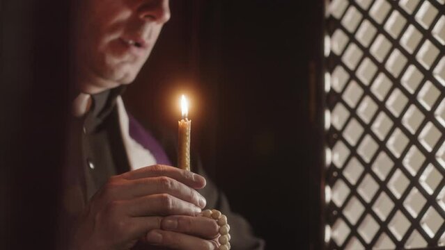 Slowmo of mature priest wearing black and purple robe blowing out candle while sitting in dark confession booth of Catholic church