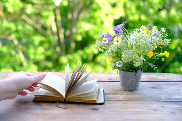 Naklejka na ściany i meble female hand hold open book in black cover, bouquet of wild flowers on wooden table in garden, blurred natural landscape in background with green foliage, concept education, knowledge