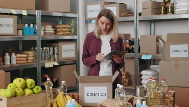 Focused food bank worker using digital tablet in hands while preparing donation boxes at warehouse. Female volunteer checking and sorting provision at charitable foundation.