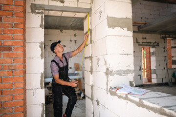 Construction worker at construction site measures the length of window opening and brick wall with tape measure. Cottage are made of porous concrete blocks, work clothes - jumpsuit and baseball cap